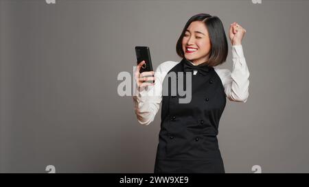 Der asiatische Kellner war zufrieden, nachdem er gute Nachrichten am Telefon gelesen hatte und viele Gäste reserviert hatten. Kellnerin mit Schürze, die mit dem Smartphone Tischreservierungen sieht, arbeitet gerne. Kamera A.. Stockfoto