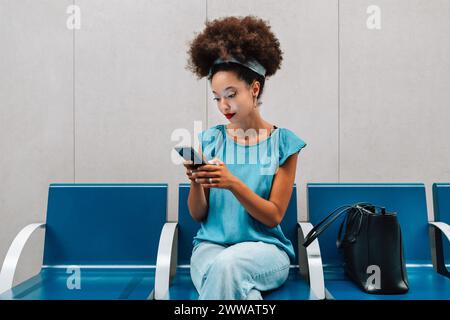 Konzentrierte junge marokkanische Frau mit Afro-Haaren und Stirnband, die auf den Bildschirm des Mobiltelefons blickt, während sie mit Handtasche im Busbahnhof unter Licht sitzt Stockfoto