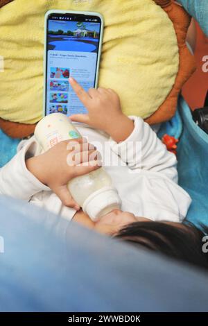 Kleines Kind in der Wiege mit seiner Flasche vor einem Telefonbildschirm. Stockfoto