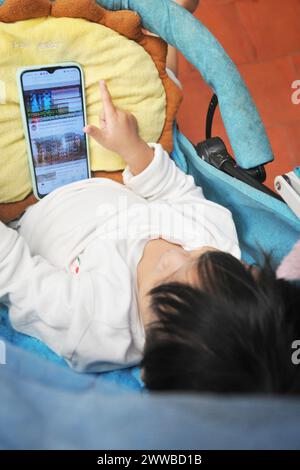 Kleines Kind in der Wiege mit seiner Flasche vor einem Telefonbildschirm. Stockfoto