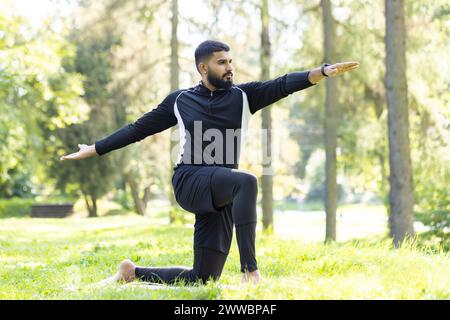 Junger Mann in schwarzer Sportkleidung, der an einem sonnigen Tag in einem friedlichen grünen Park eine Yoga-Pose spielt. Stockfoto