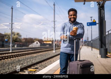 Ein glücklicher Mann mit Koffer trinkt gern Kaffee, während er auf dem Bahnhof steht. Stockfoto