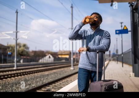 Ein glücklicher Mann mit Koffer trinkt gern Kaffee, während er auf dem Bahnhof steht. Stockfoto