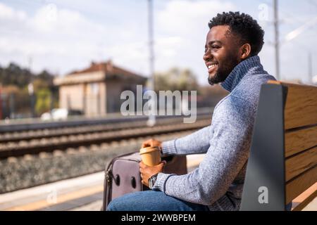 Glücklicher Mann mit Koffer trinkt Kaffee, während er auf einer Bank am Bahnhof sitzt. Stockfoto