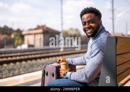 Glücklicher Mann mit Koffer trinkt Kaffee, während er auf einer Bank am Bahnhof sitzt. Stockfoto