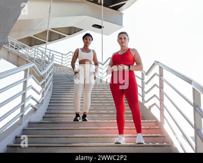 Zwei Frauen in Fitnesskleidung mit verschiedenen Farben, die auf einer Treppe in die Kamera schauen. Eine Frau in Übergröße mit ihrem Fitness-Freund posiert Stockfoto