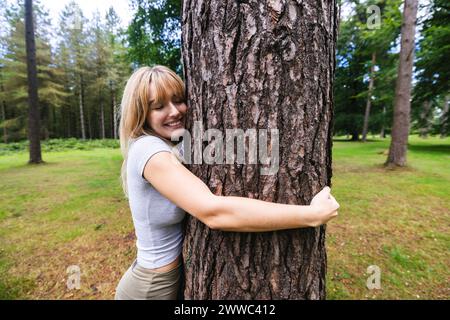 Lächelnde junge Frau, die im Wald Baumstamm umarmt Stockfoto