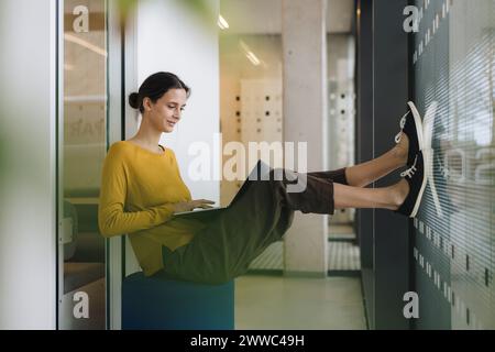 Lächelnde junge Geschäftsfrau, die im Bürokorridor sitzt und einen Laptop benutzt Stockfoto