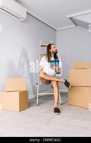 Junger Mann, der auf einer Leiter mit Tablet-PC in der Nähe von Kartonschachteln sitzt Stockfoto