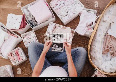 Schwangere Frau, die auf dem Boden sitzt und zu Hause Ultraschallbilder hält Stockfoto