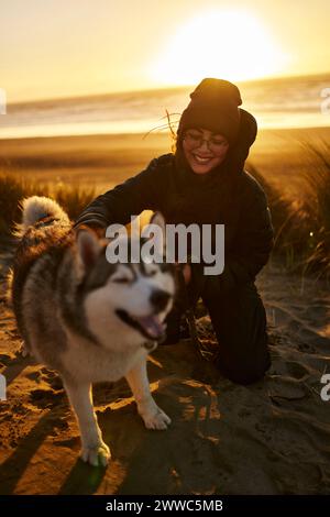 Lächelnde Frau kniend und streichelt Husky-Hund am Strand Stockfoto