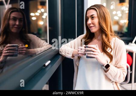 Lächelnde Geschäftsfrau mit Kaffeetasse in der Nähe des Fensters im Büro Stockfoto