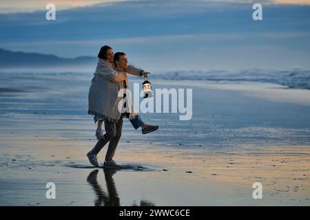 Junger Mann Huckepack Frau mit Laterne am Ozean Strand bei Sonnenuntergang Stockfoto