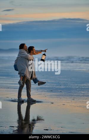 Mann Huckepack Frau mit Laterne, die auf das Meer am Strand zeigt Stockfoto