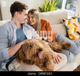 Glückliches junges Paar, das zu Hause mit flauschigem Hund auf dem Sofa sitzt Stockfoto