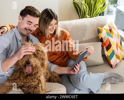 Glückliche junge Frau mit Tablet-PC und Mann, der Hund zu Hause streichelt Stockfoto