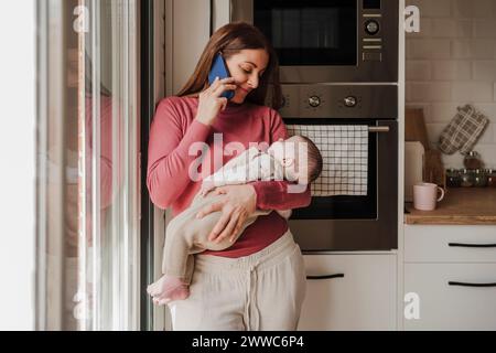Lächelnde Frau, die mit dem Smartphone spricht und die Tochter in der Küche trägt Stockfoto