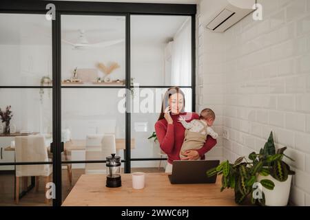 Glückliche freiberufliche Mutter, die am Smartphone spricht und ein Mädchen in der Küche trägt Stockfoto
