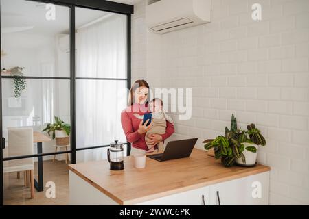 Freischaffende Mutter, die ihr Smartphone benutzt und ihr Baby in der Küche trägt Stockfoto