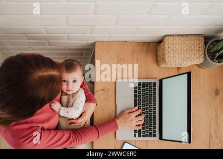 Freiberuflich tätige Mutter tippt auf dem Laptop und hält das Baby fest Stockfoto