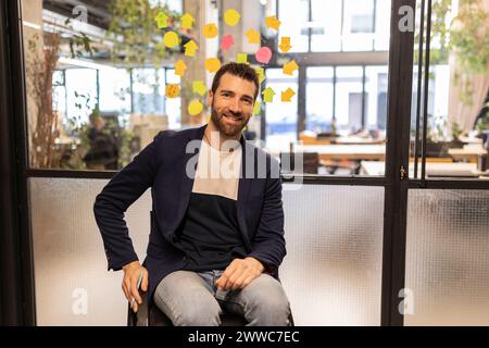 Lächelnder Geschäftsmann, der im Rollstuhl neben der Glaswand im Büro sitzt Stockfoto