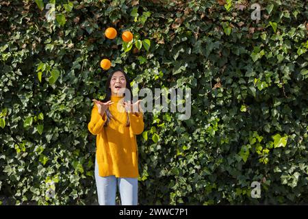 Glückliche junge Frau, die Orangen vor Pflanzen jongliert Stockfoto