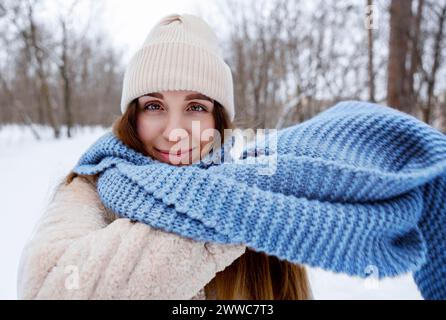 Lächelnde Frau mit Strickmütze und blauem Schal im Winterwald Stockfoto