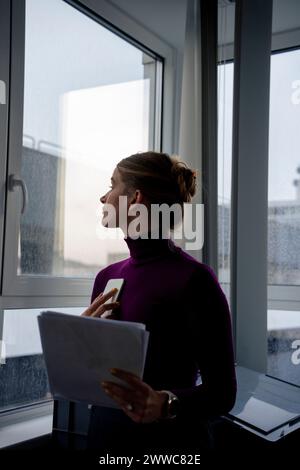 Geschäftsfrau, die Dokumente hält und im Büro aus dem Fenster blickt Stockfoto