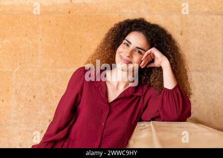 Lächelnde Frau mit lockigem Haar, die sich vor der Wand am Ellenbogen beugt Stockfoto