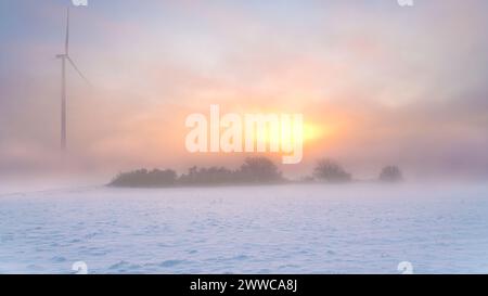 Deutschland, Hessen, Hunfelden, Schnee vor kleinem Hain und Windturbine bei nebeliger Wintersonnenaufgang Stockfoto