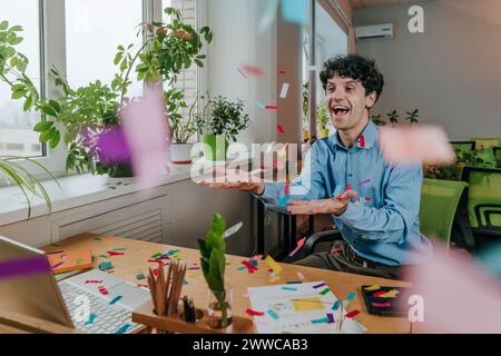 Aufgeregter Geschäftsmann, der am Schreibtisch im Büro mit Konfetti spielt Stockfoto