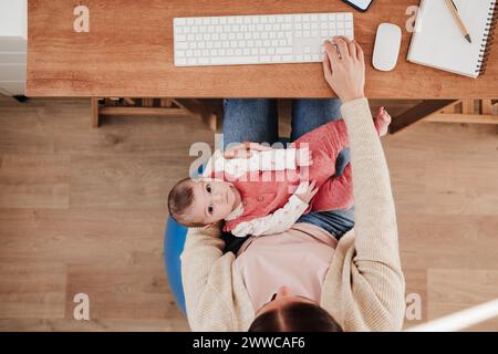 Niedliches Mädchen, das auf dem Schoß der arbeitenden Mutter im Heimbüro liegt Stockfoto
