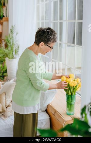 Reife Frau arrangiert Tulpen in Vase auf Fensterbank zu Hause Stockfoto