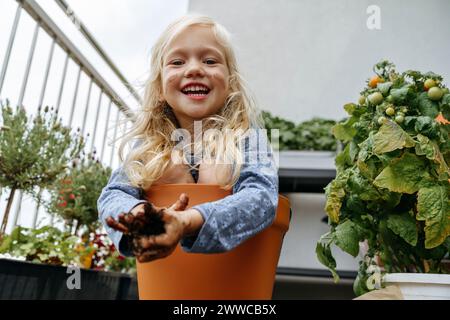 Fröhliches Mädchen mit schmutzigen Händen, das im Topf neben Pflanzen auf dem Balkon sitzt Stockfoto