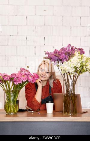 Lächelnder Blumenhändler, der sich neben verschiedenen Blumen auf dem Schreibtisch lehnt Stockfoto