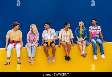 Fröhliche Freunde mit bunten Kleidern sitzen zusammen auf der gelben Wand Stockfoto