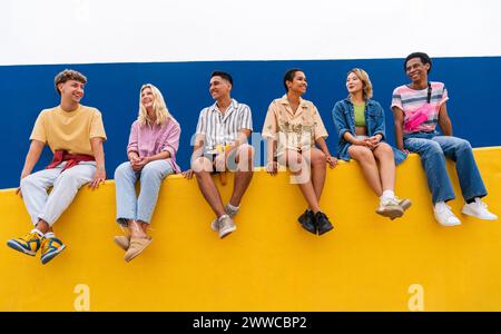 Fröhliche Freunde mit bunten Kleidern sitzen zusammen auf der gelben Wand Stockfoto