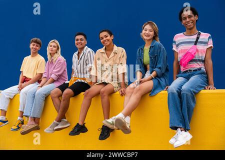 Lächelnde Freunde mit bunten Kleidern sitzen zusammen auf der gelben Wand Stockfoto