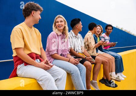 Glückliche Freunde mit bunten Kleidern, die auf der gelben Wand sitzen und Smartphones halten Stockfoto