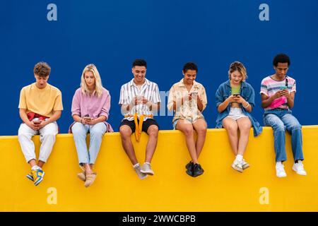 Fröhliche Freunde mit bunten Kleidern, die auf Gelb sitzen, mit ihren Smartphones Stockfoto