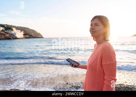 Nachdenkliche Frau mit Smartphone am Strand Stockfoto