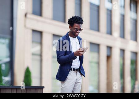 Stilvoller afroamerikanischer Mann in eleganter Freizeitkleidung, mit Mobiltelefon vor dem Hintergrund der Stadt. Modischer Kerl mit Smartphone im Freien. Stockfoto