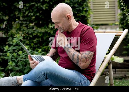 Reifer Freiberufler mit Hand am Kinn mit Laptop im Garten Stockfoto