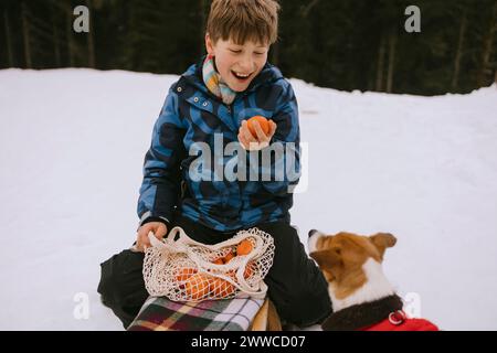 Glücklicher Junge, der Orange in der Nähe des Hundes im Winterwald hält Stockfoto