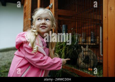 Blondes Mädchen mit rosafarbenem Regenmantel, das Kaninchen im Käfig füttert Stockfoto