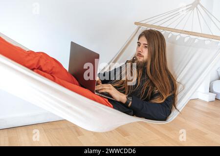 Junger Programmierer auf Hängematte mit Laptop zur Programmierung beim Start im Büro Stockfoto