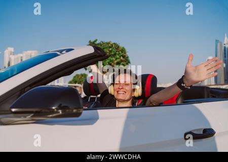 Glückliche Frau, die kabellose Kopfhörer trägt und die Arme in einem weißen Cabriolet am sonnigen Tag gestreckt hat Stockfoto