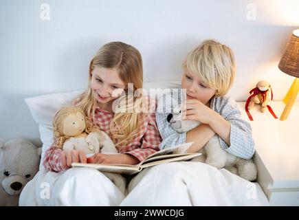 Ein lächelndes Mädchen liest ein Buch mit einem Bruder, der zu Hause auf dem Bett sitzt Stockfoto