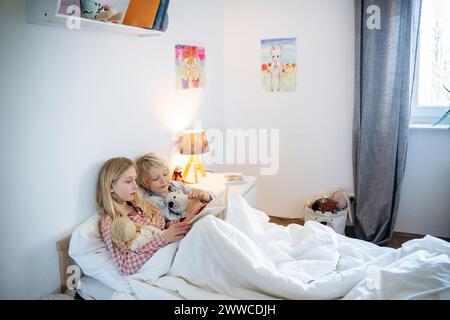 Der blonde Bruder und die blonde Schwester lesen zu Hause ein Buch im Bett Stockfoto