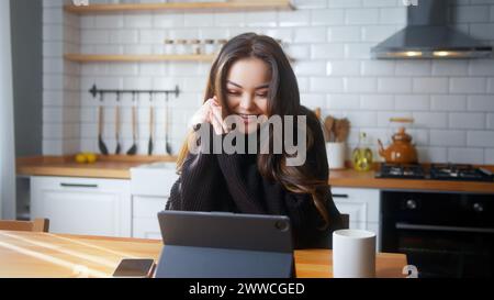 Lächelnde junge asiatische Frau sitzt in der Küche und führt Videogespräche über einen Tablet-Computer. Teilen Sie Nachrichten, sprechen Sie mit der Familie, genießen Sie Webchat virtuell Stockfoto